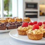 Plates of breakfast pastries including chocolate muffins and oatmeal muffins topped with fresh raspberries, displayed on a bright kitchen counter.